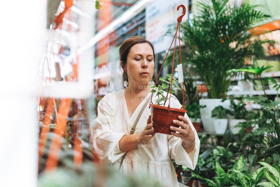 Brunette middle aged woman in white dress buys green potted house plants at the garden store