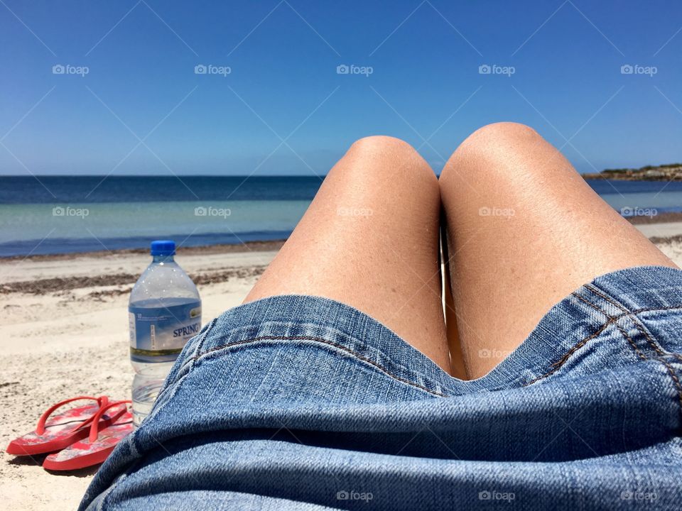 Leg shot, woman with tanned bare legs in denim
Skirt laying on remote secluded beach water bottle by her side south Australia 