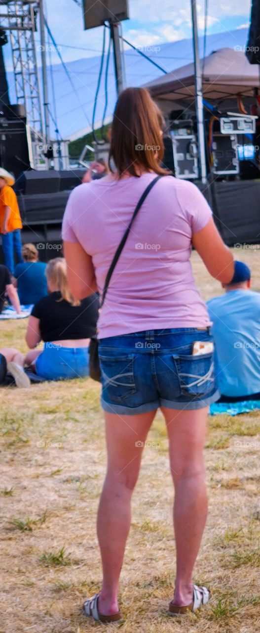 woman wearing a pink shirt, denim shorts, and a black purse waiting for a concert at an Oregon fair in Summer