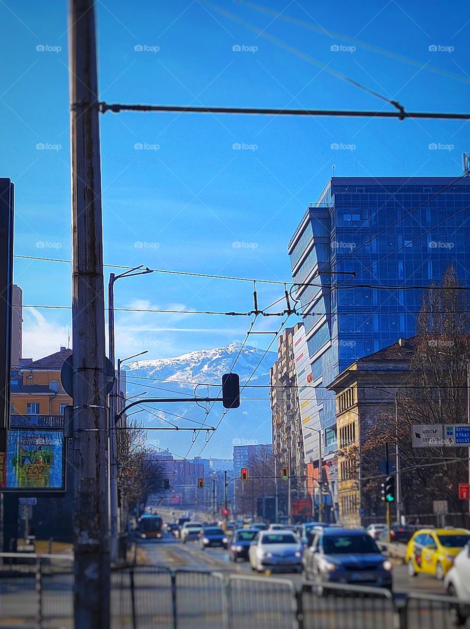 A photo of a busy Boulevard in the capital of Bulgaria with many cars, buildings, light poles and a beautiful snowy mountain in the distance