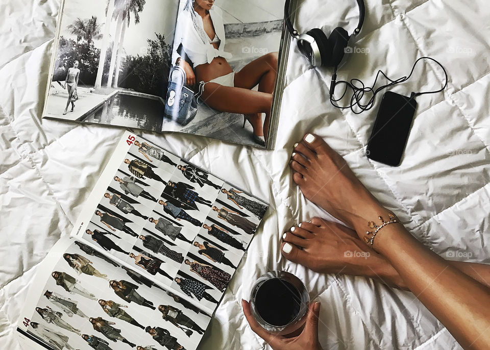 Young woman reading modern magazines, enjoying wine and listening to the music from her mobile phone in cozy bed 