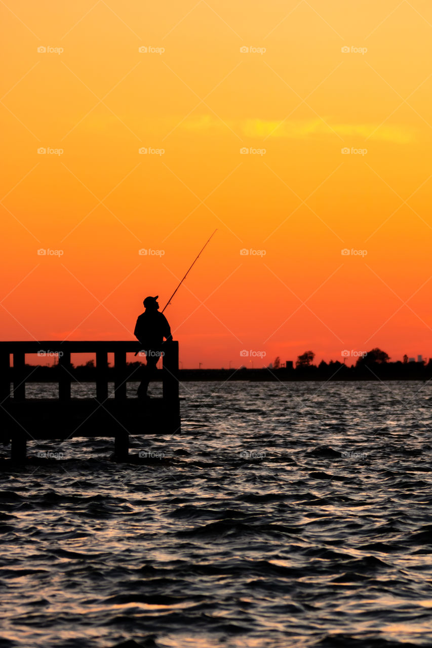 Silhouette of a fisherman standing on a pier, looking out across the sea as the summer sky burns a deep orange at sunset 