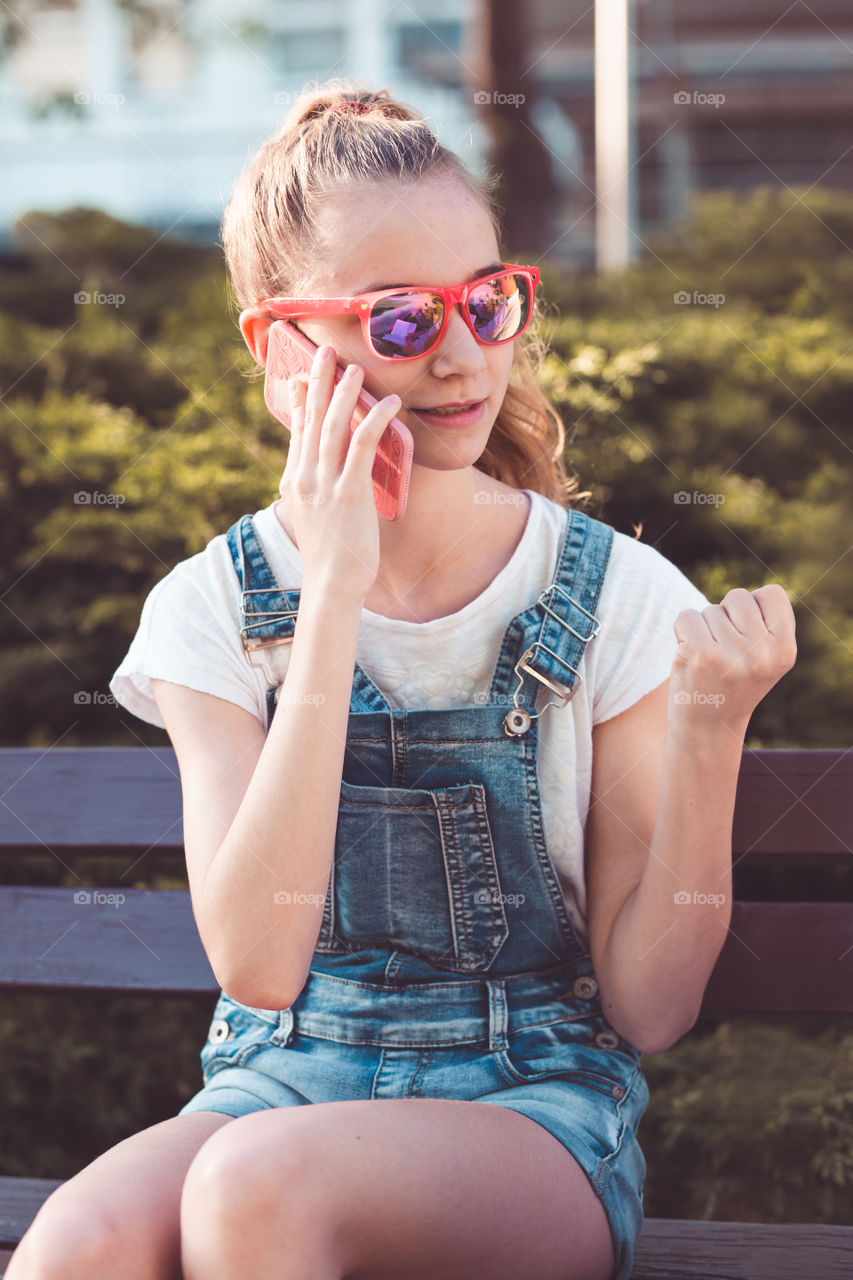 Young girl talking on the mobile phone wearing blue jeans and sunglasses sitting on a bench in a park