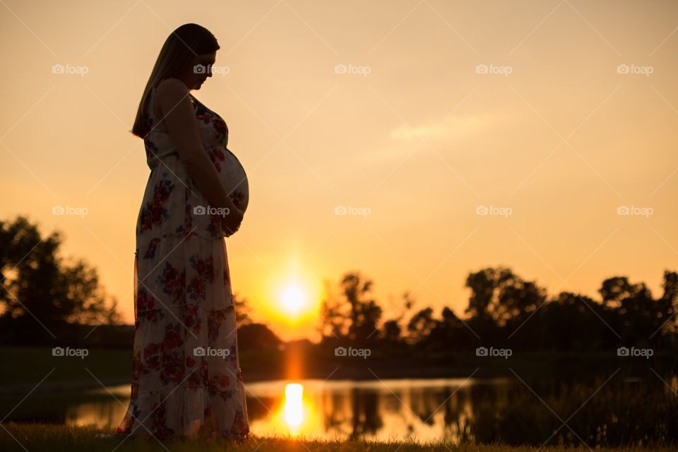 Pregnant woman holding belly during an orange sunset on the lake. Sun reflecting on the water. 