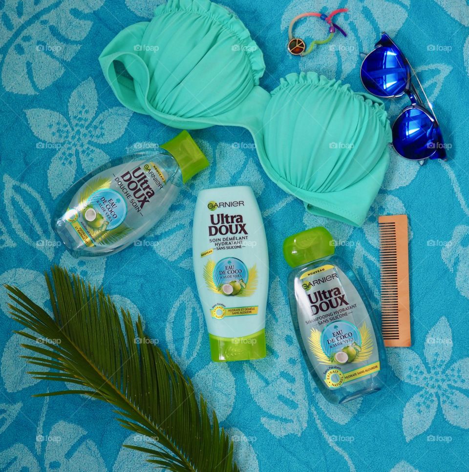 Garnier eau de coco shampoo, conditioner and bath gel on a blue towel with comb and palm leaf and blue sunglasses and bikini.