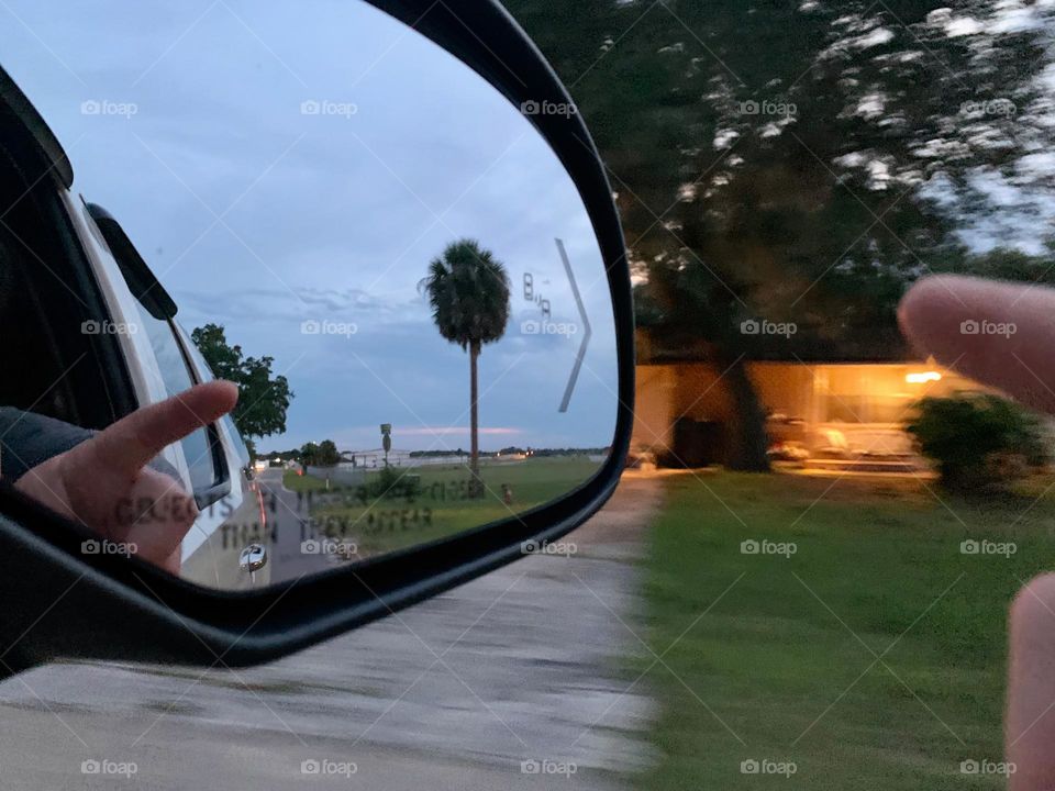 Commuting: passenger mirror on a vehicle where seeing something the passenger points at by a private airport with palm tree and trees and house with lights on during the evening on a cloudy day.