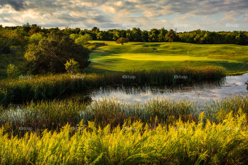 A lush green golf course bathed in the golden glow of the morning sun. The green is glowing as the warm sunlight peeks out from behind the low morning clouds. If you look closely, you can see 2 well placed shots next to the flag.