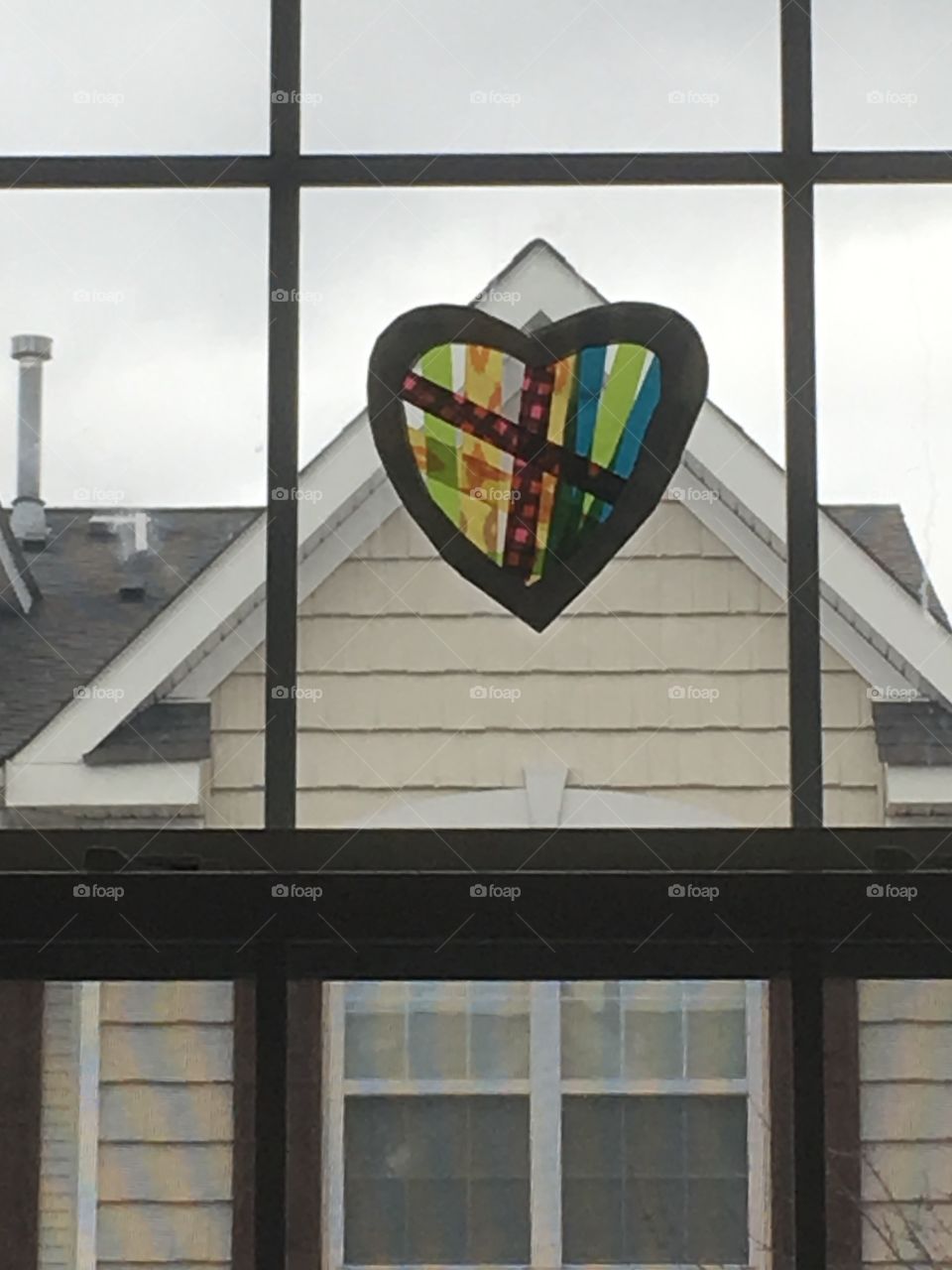Home is where the heart is. Child's heart artwork hung in window. Peaked roof of house seen through window. 