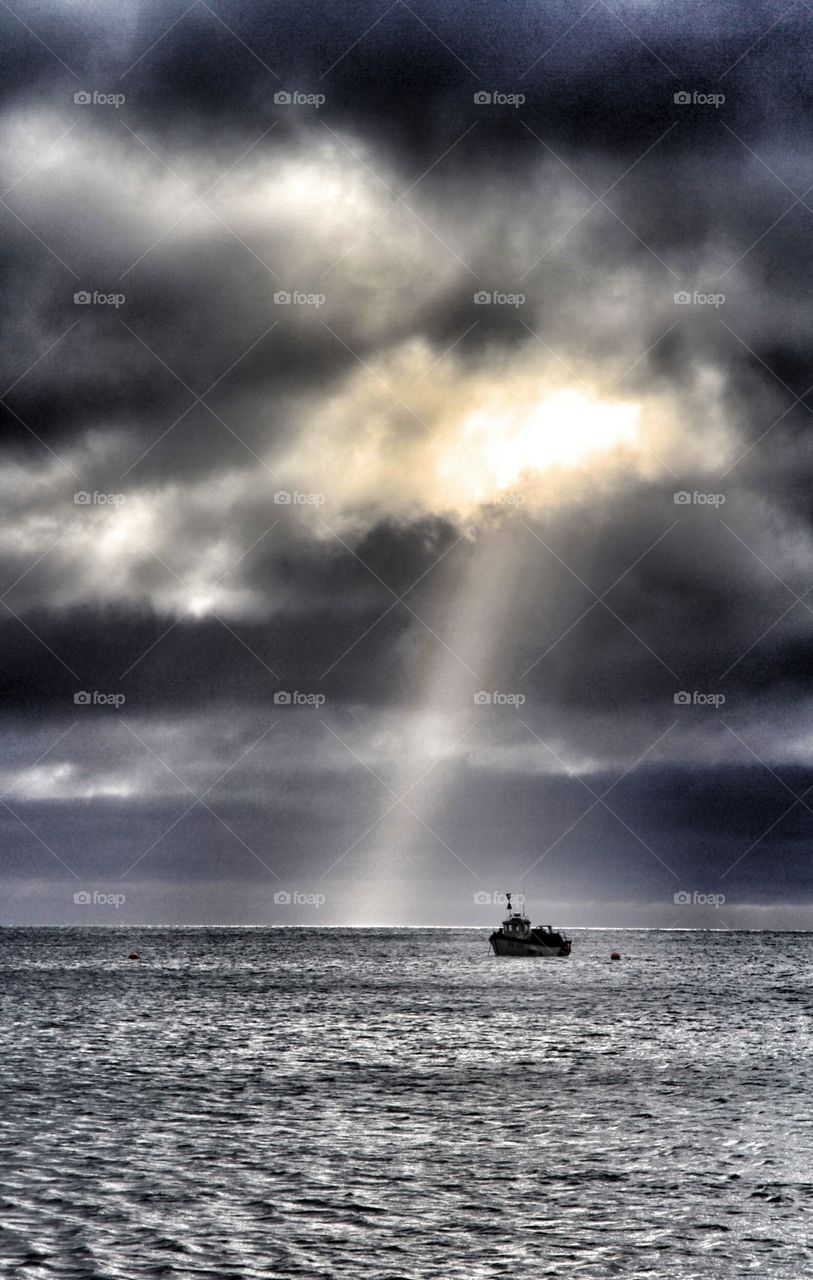 Sun Ray Gun. A bright shaft of sunlight breaks through a stormy sky over a dark sea with a lone fishing boat beneath.