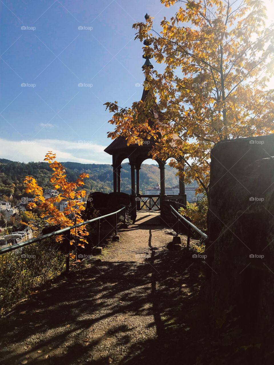 In the middle is a dirt road , the two sides are fenced with iron bars , trees with beautiful yellow leaves .the sun shines through the leaves, big rock on the right.And standing here can see the surrounding landscape very beautiful