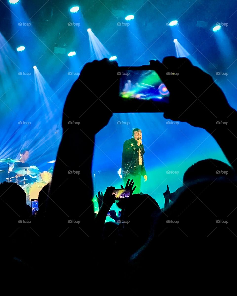 Photo of a band playing seem through the silhouette of a fan taking a video in their phone