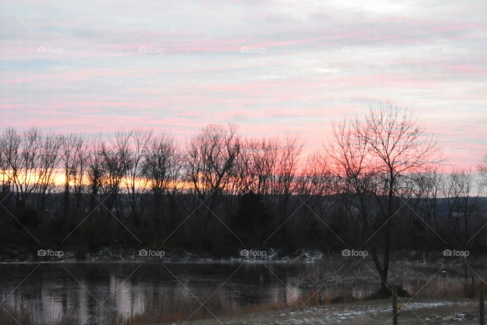 Beautiful, pastel colored sunset. There's a pond with tree silhouettes behind it and spots of snow on the ground.