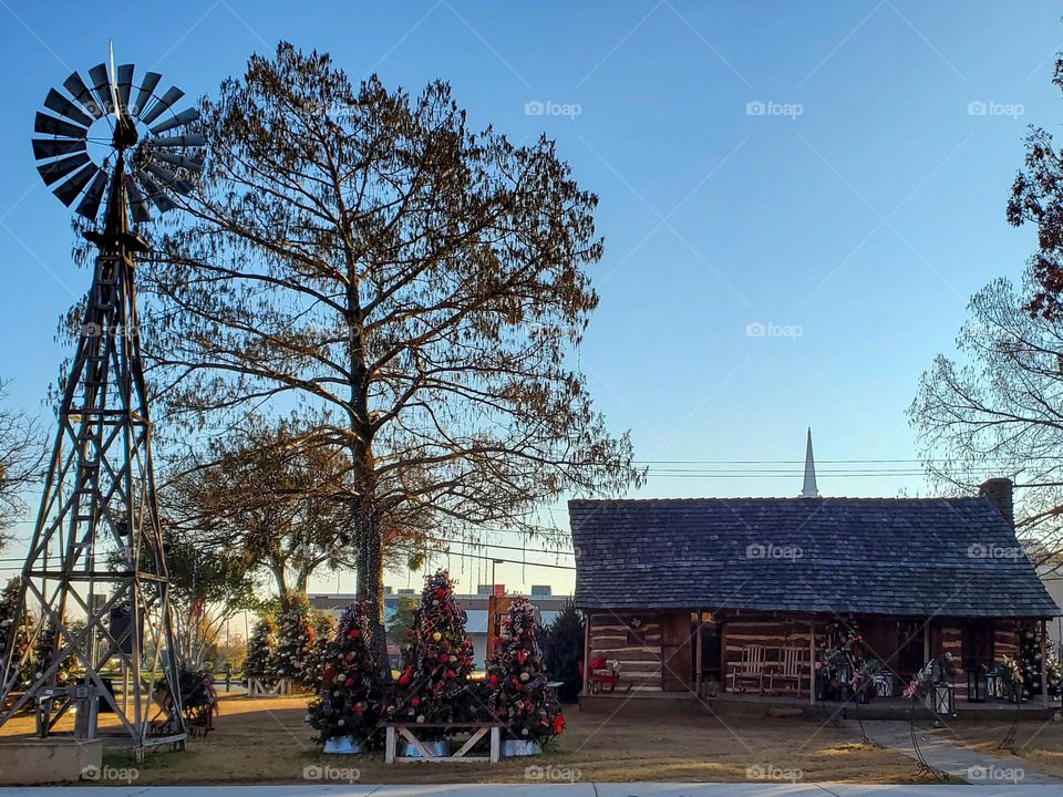 A windmill sits next to a log cabin that is decorated for the Christmas season