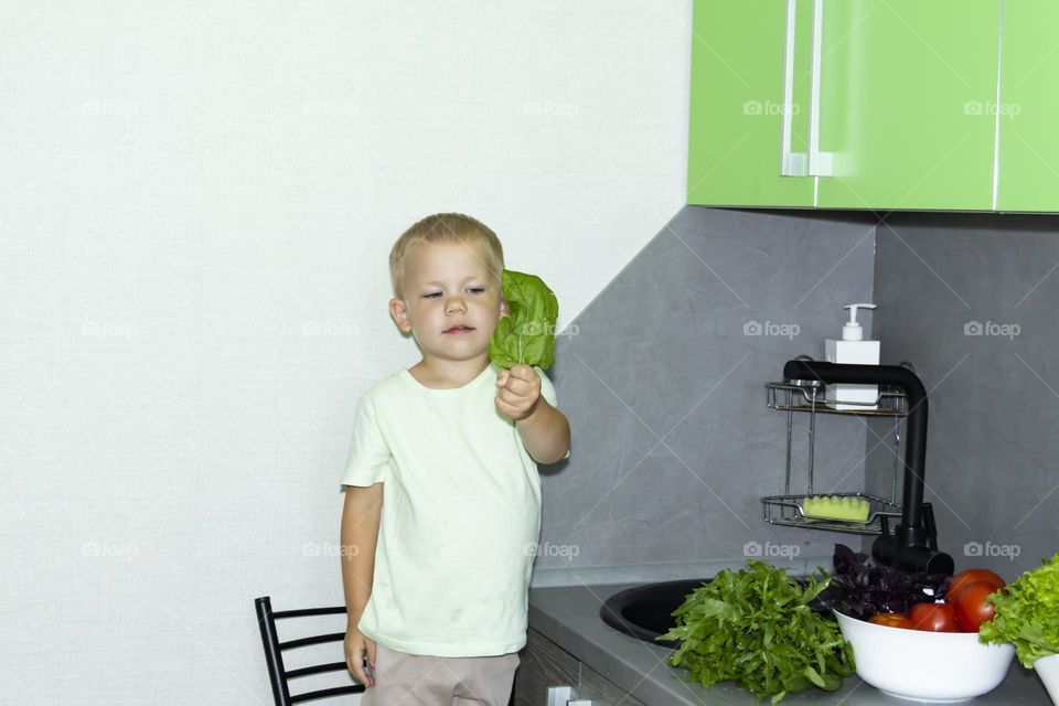 A small child in the kitchen near the sink with spinach in his hands.
