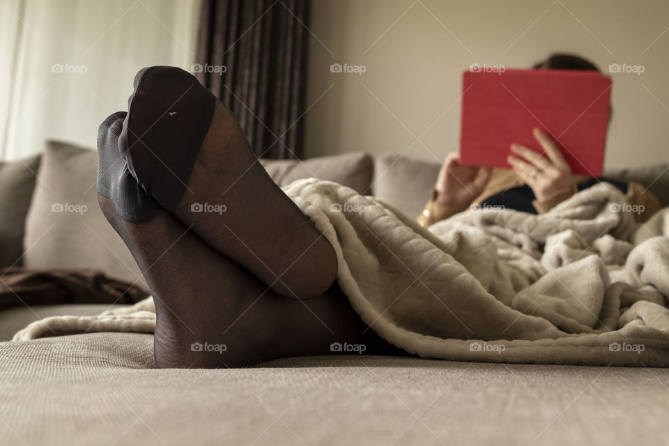 A portrait of the pantyhosed feet of a woman resting in a couch and browsing on her tablet. the pantyhose are black and dhr also has a cosy blanket pulled over her.