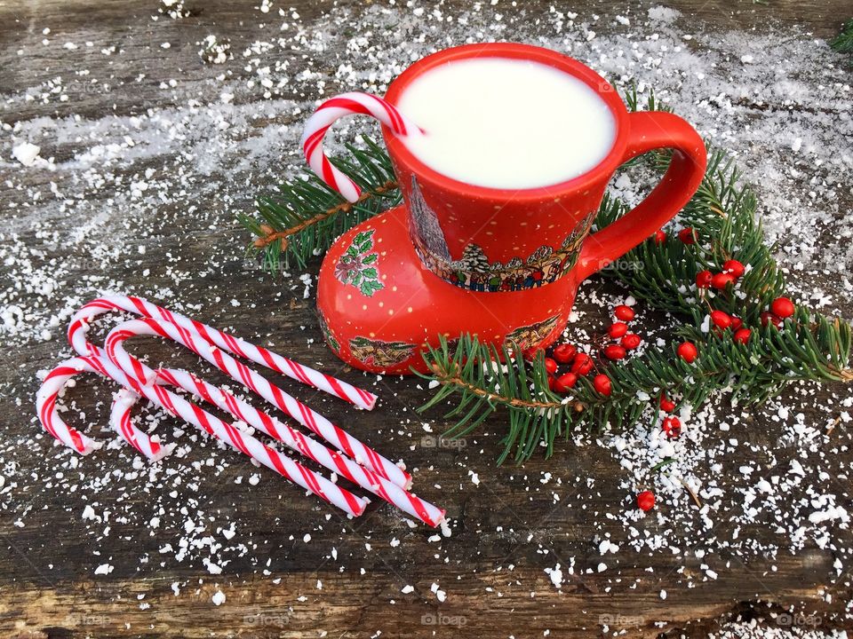 Boot shaped mug filled with milk and candy canes with candy canes and pine cone tree branches on snowy table 