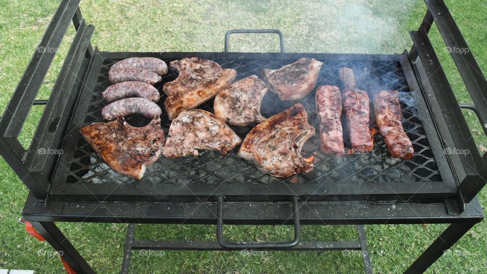 Backyard barbecue and Braai. Man braai barbecue sausages, food meat at home in the yard. Men grilling lots of meat on steel grill. Braai steak pork chops sausages and braai bacon. Cooking at home and outdoor cooking. Make a hotdog in bun bread roll.