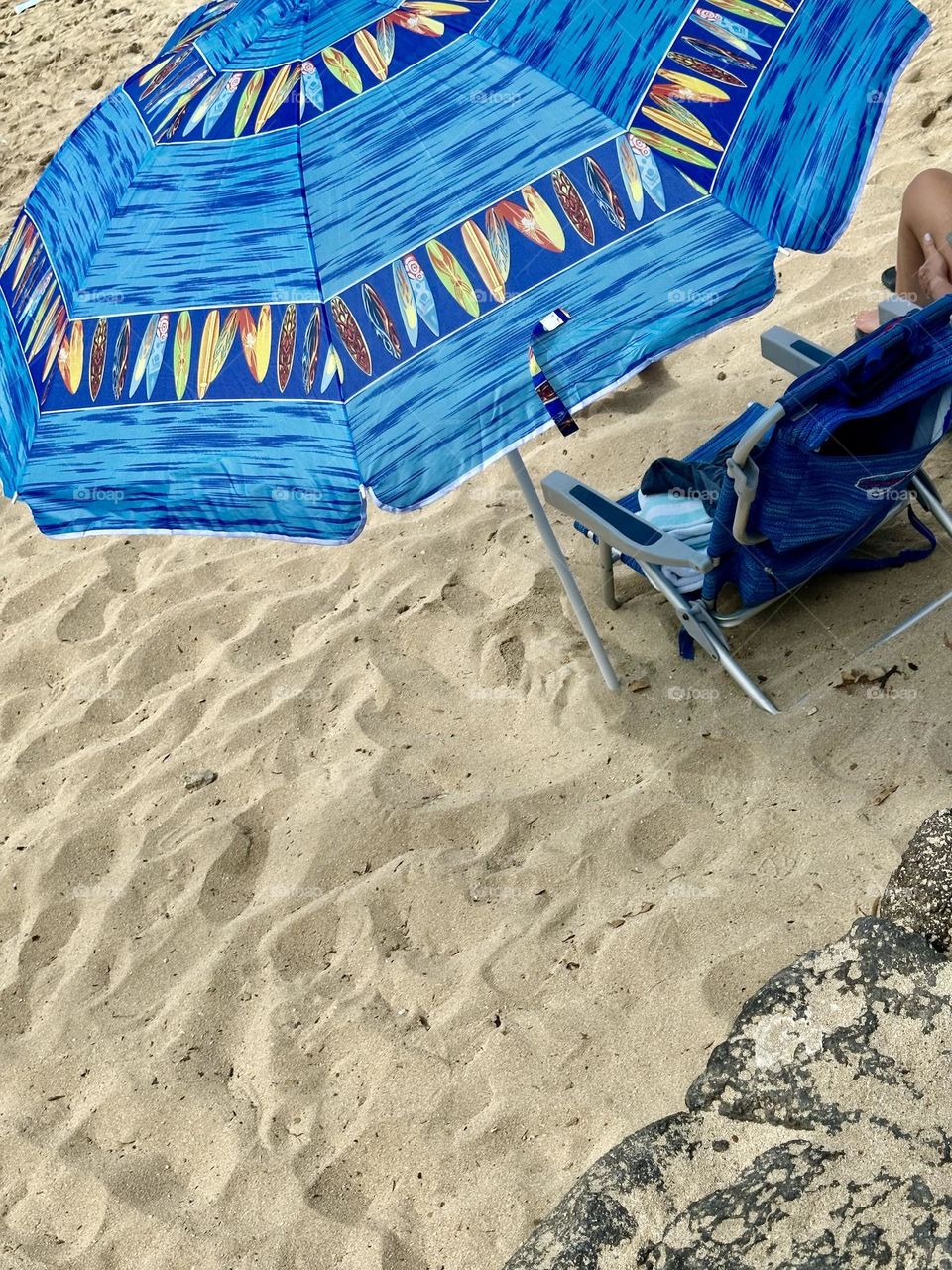 Blue beach umbrella with surfboard design and serious canvas beach chairs, one occupied, set up in the sand 