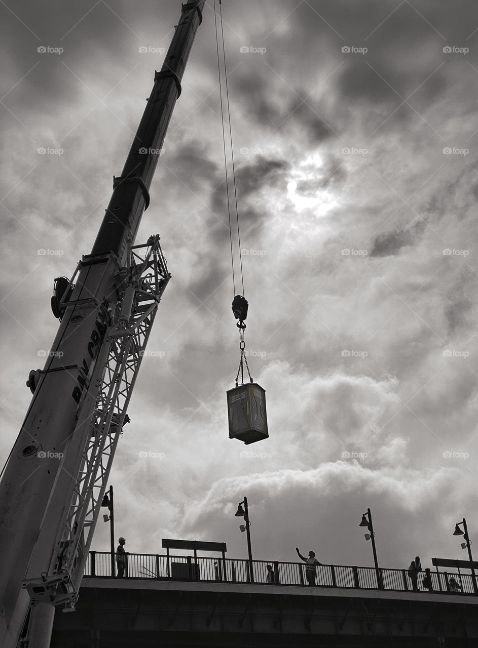 Crane delivering a portable toilet to workers on an elevated train platform 