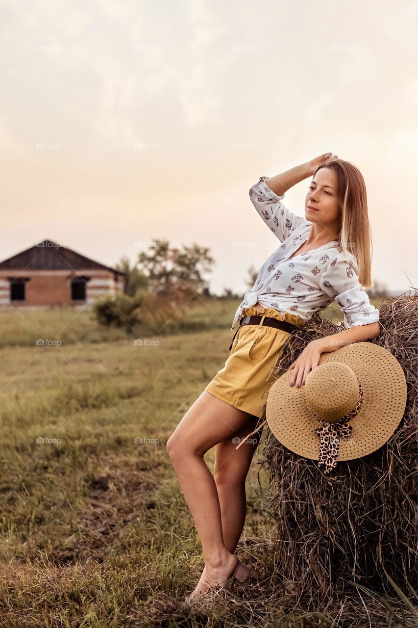 Young blonde girl in a field with haystacks, wearing a straw hat and smiling. Country life. Life style portrait