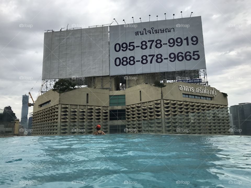 A woman in orange cap swimming in an infinity pool in bangkok. In the background there is a large dirty building