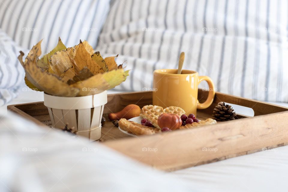 Wooden tray with a yellow mug of coffee, waffles in a saucer and a bouquet of autumn poplar leaves in a wicker basket lying on a straightened bed, close-up side view. Concept breakfast in bed.