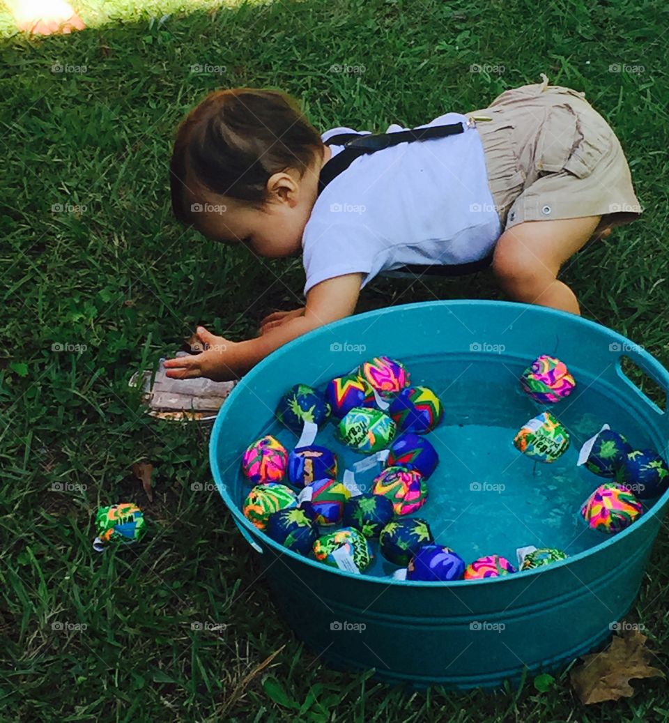 One year old at his birthday party picking up a little toy from the ground next to water in tiny tub. 