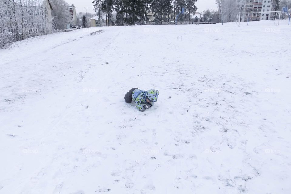 A small, carefree boy walks in winter through the white snow in the park, near the trees in the snow.