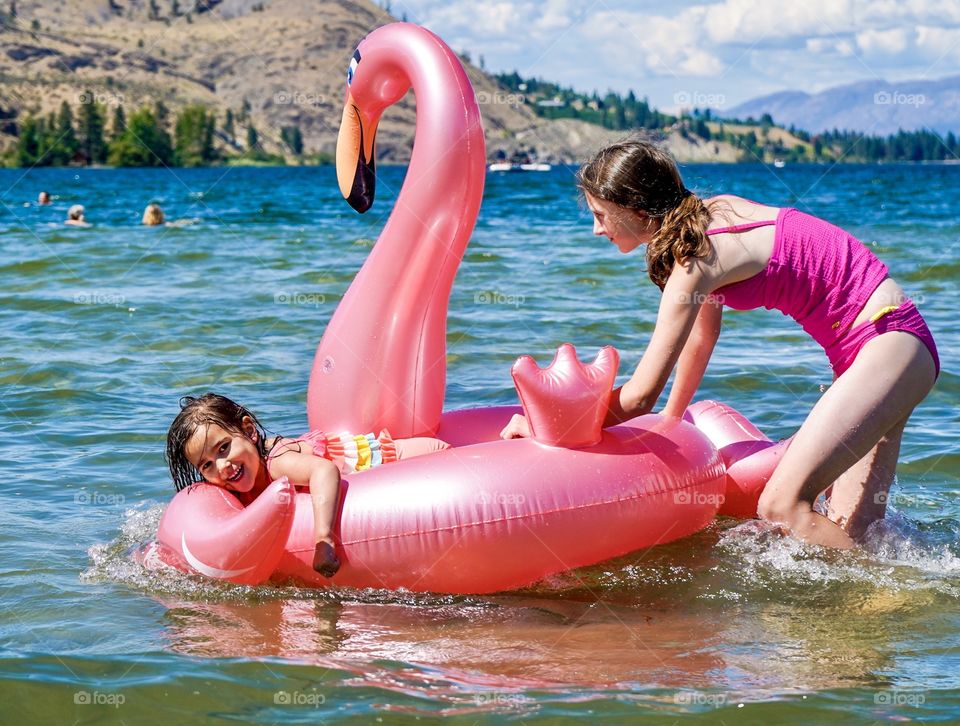 Kids playing on pink flamingo toy on a sunny lake in summer 
