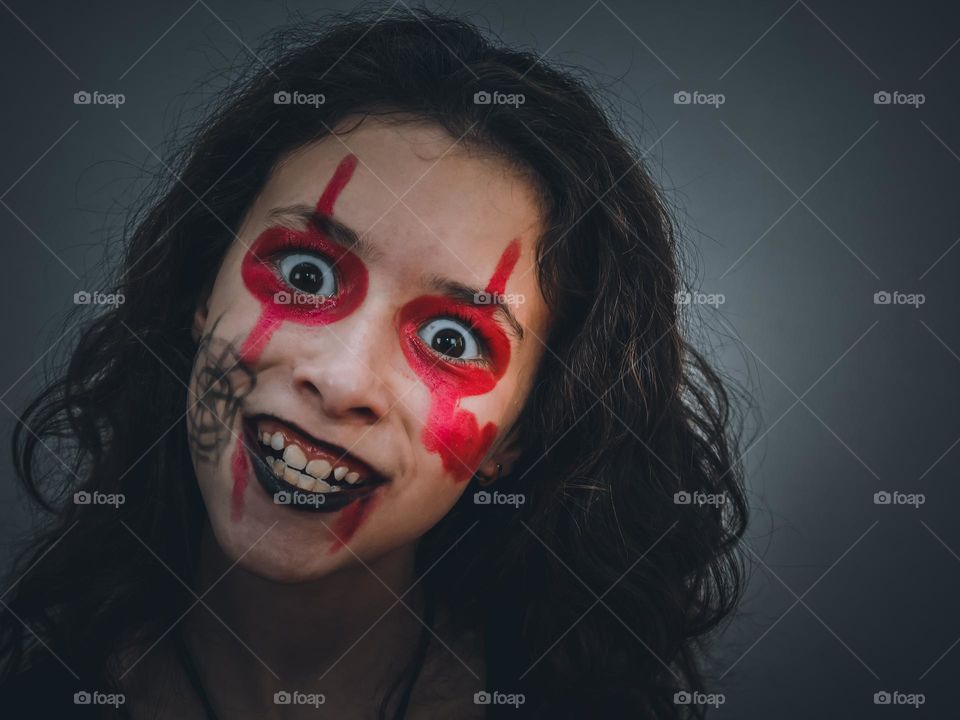 Portrait of a beautiful caucasian teenager girl with shoulder-length wavy brown hair with red-black halloween make-up on her face expresses a frightening angry emotion, close-up side view. Halloween concept.