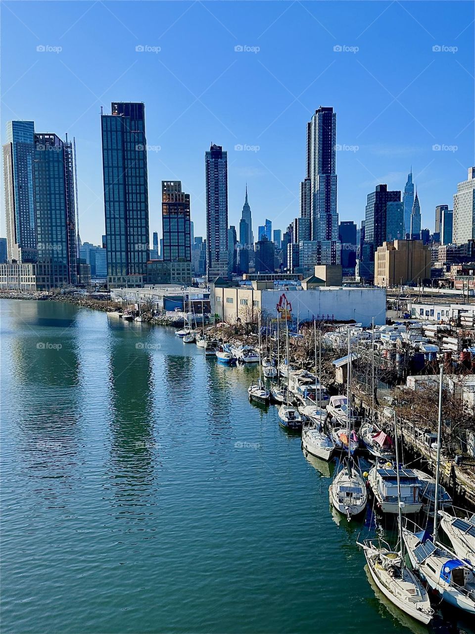 This is picturesque „Newtown Creek“ with its many boats seen from the „Pulaski Bridge“ that connects „Greenpoint“, Brooklyn to LIC, Queens. In the distance we see „Manhattan“ incl. landmarks such as the „Empire State Bldg“. 2024. Hypnotic Productions