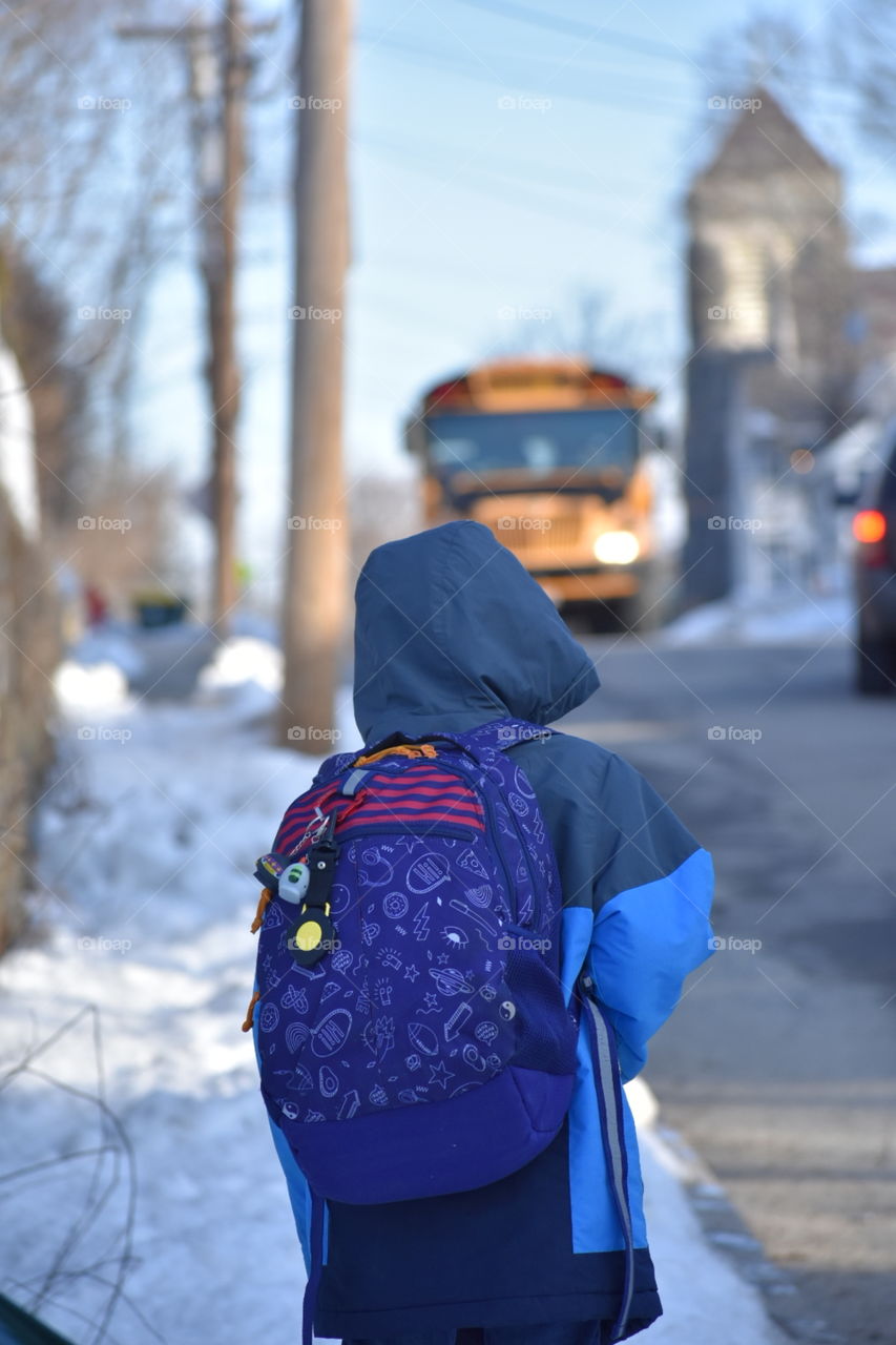 little boy in warm blue jacket standing outside in the snow, waiting for the bus to arrive. bus is up the street. snow is on the sidewalk.