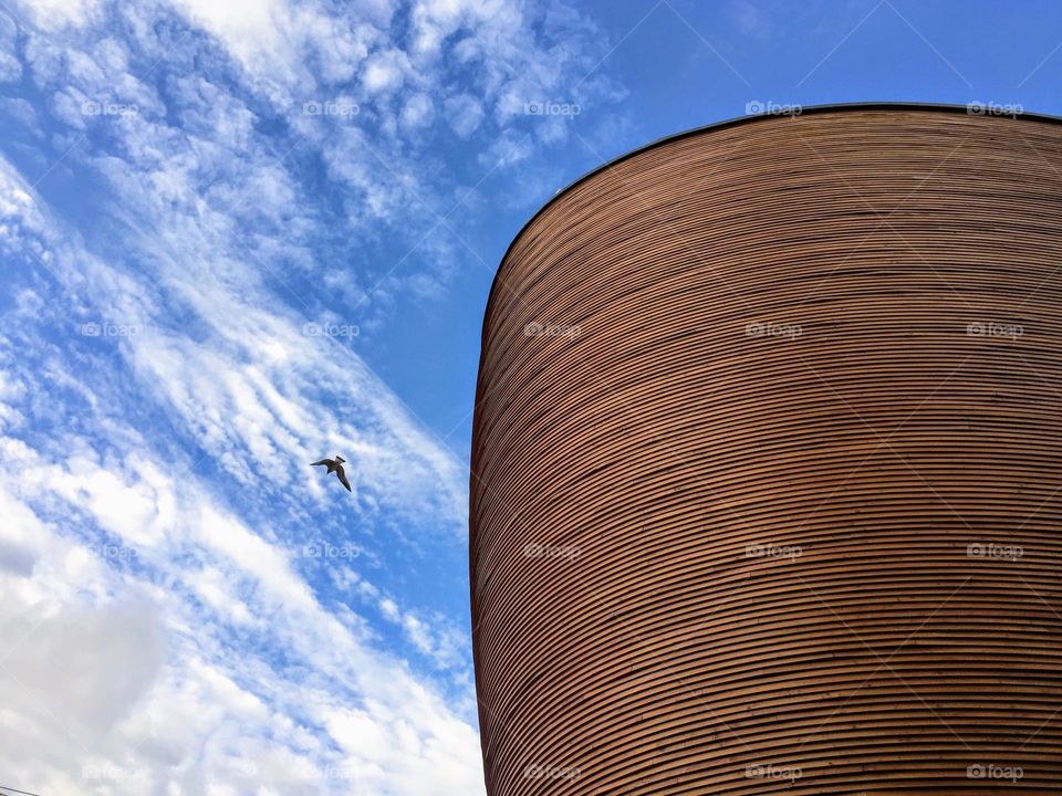 Unusual angle view up to the Kamppi modern wooden round oval chapel of silence against the deep blue sky with white clouds and flying seagull 