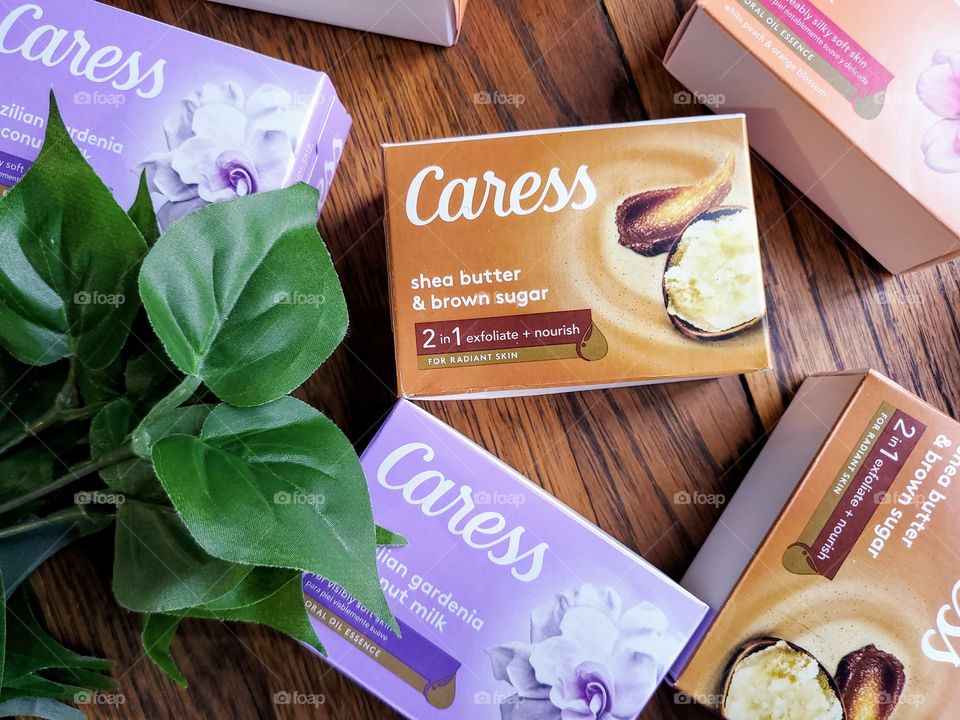 The new look Caress bar single packs: Daily silk, shea butter & brown sugar, and Brazilian gardenia & coconut milk on a brown wood surface with accenting green leaves on the left.