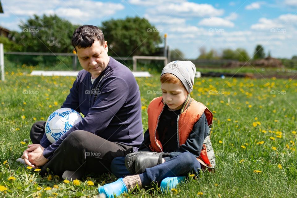 Father and son have fun together, sit on the grass in the summer in the village after playing ball football, lifestyle style
