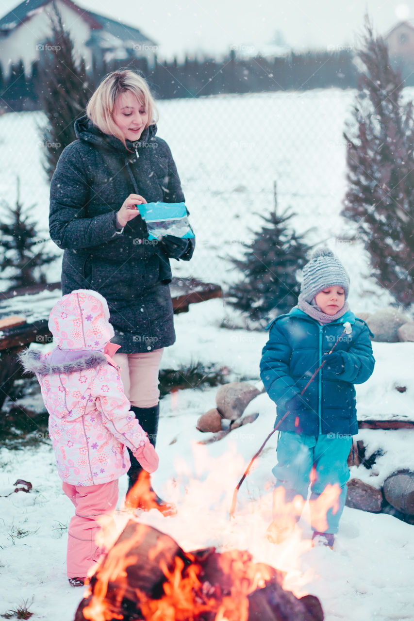 Family spending time together outdoors in the winter. Parents with children gathered around the campfire preparing marshmallows and snacks to toasting over the campfire using wooden sticks