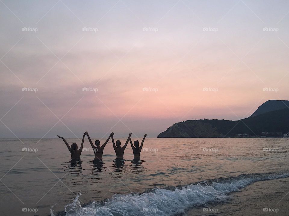 Four girls in the sea water with their hands up in a colorful sunset and a mountain in the background