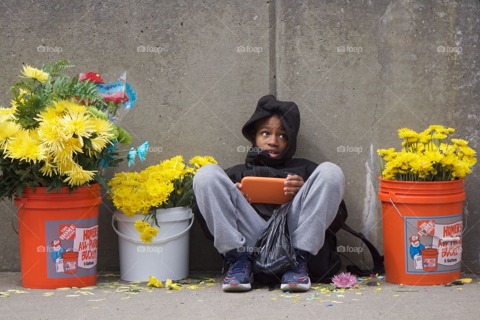 An urban street scene in The Bronx, New York City as a boy sits on the sidewalk between buckets of colorful flowers waiting for a local parade to begin.