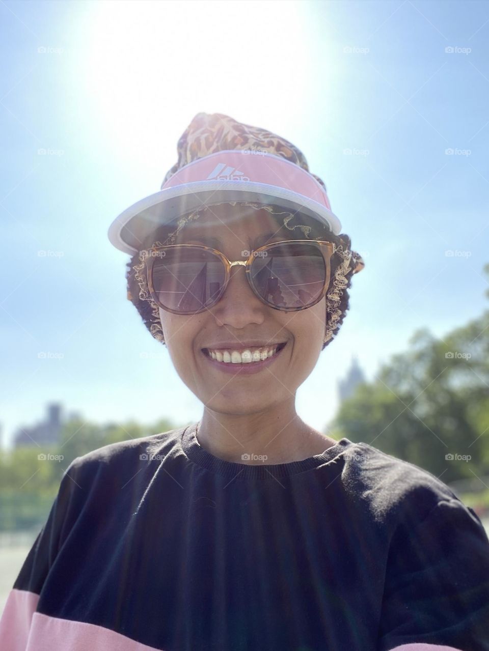 Portrait of a woman smiling wearing a leopard hair bonnet and pink hat to protect her hair from dust. With sky and trees backdrop. 