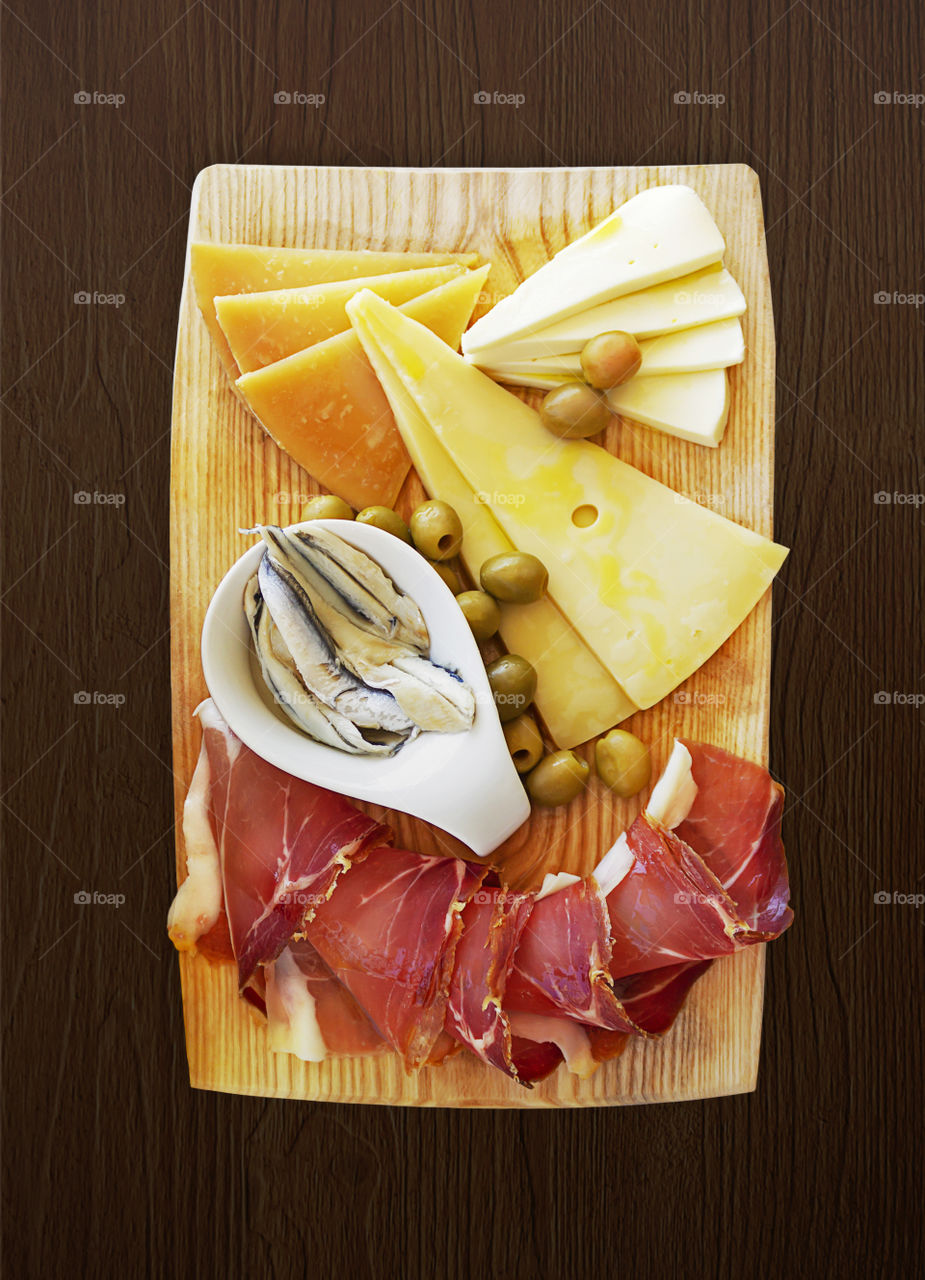 Croatian traditional food, Dalmatian plate. Ham, cheese sprinkled with olive oil with capers in a pickle with olives on a light wooden board on a dark wooden background. The perfect appetizer for wine