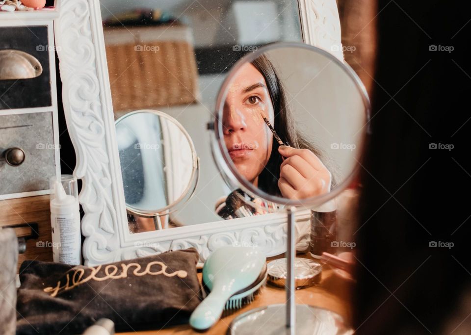 Close-up of a woman applying concealer to her face while looking into a small mirror