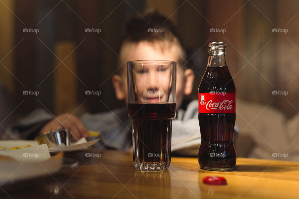 a cheerful, cute guy of fifteen years old, sitting in a cafe, smiling, having fun and looking through two bottles of Coca-Cola.  dressed in denim, charismatic teen