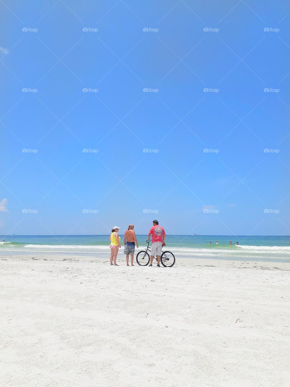Three friends chat while standing on the beach. One of the friends is holding a bicycle. They are facing the ocean and away from camera.