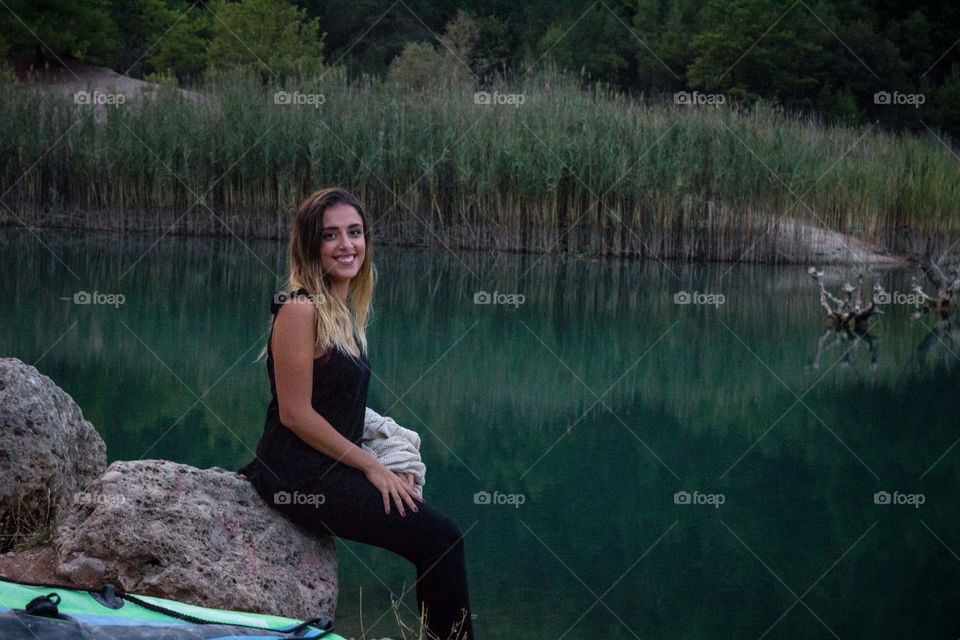 beautiful gild poses near a green lake sitting on a rock and smiling