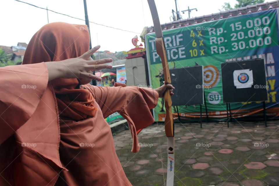 An Asian woman in a hijab holds a bow, aims and releases an arrow target at the aiming board right in front of her, Semarang, Indonesia.