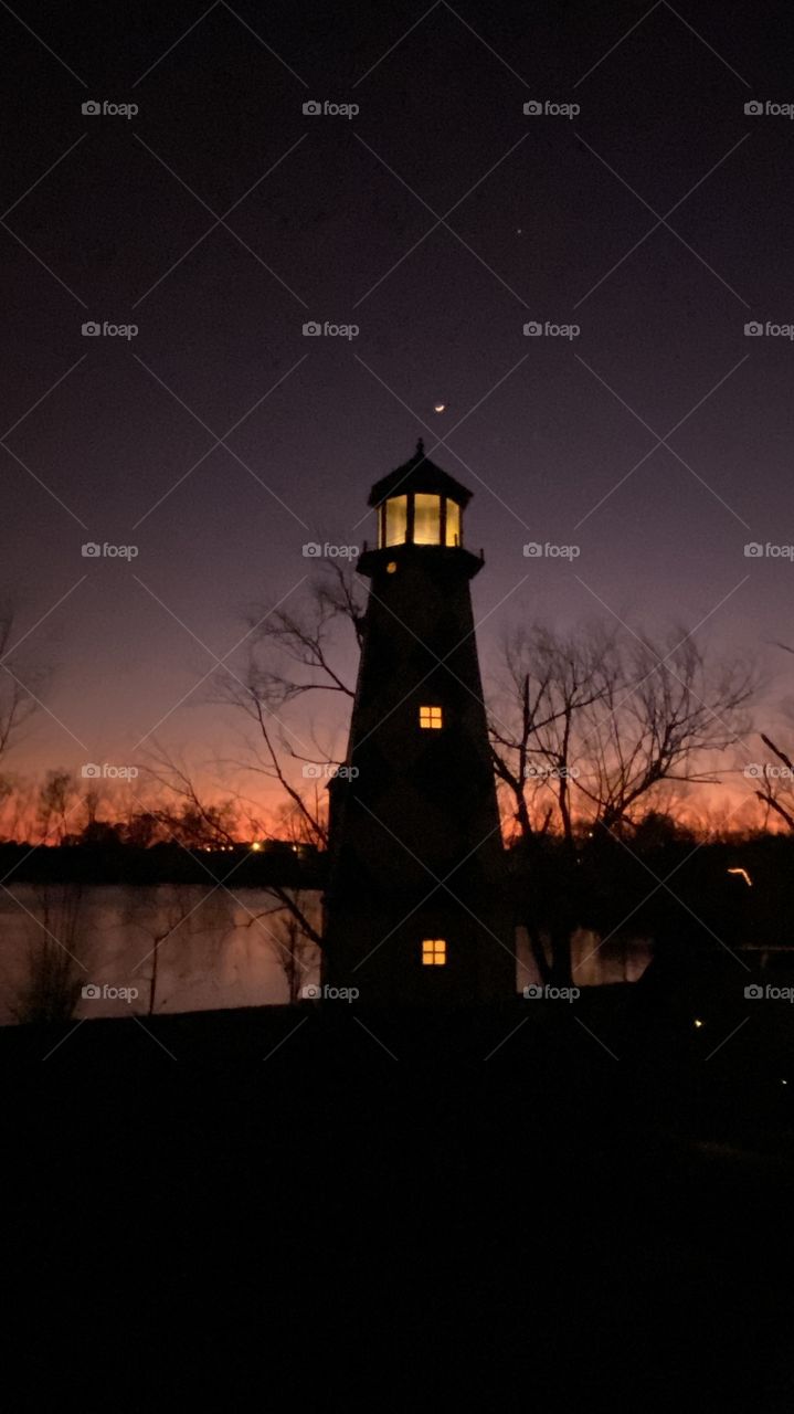 Majestic Lighthouse stands Strong against a Powerful Backlit Horizontal Glow of Orange Reds Yellow glow backing up the night Violet-Pinkish Sky. Good Night. 