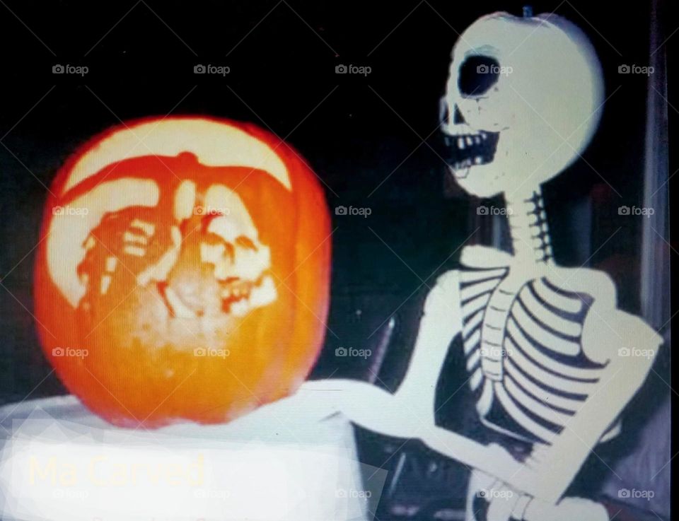 Hand carved pumpkin of Grim REAPER with sickle. Candle burning inside lights up to scare kids. Halloween decorations on porch in dark. Skeleton sits by.