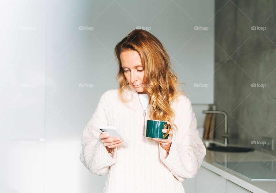 Young blonde woman in white knitted sweater with cup of tea using mobile phone in kitchen at home 