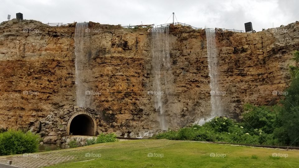 The side of a cliff with three water falls coming over the side. Train tracks come to a tunnel in the middle of the cliff.