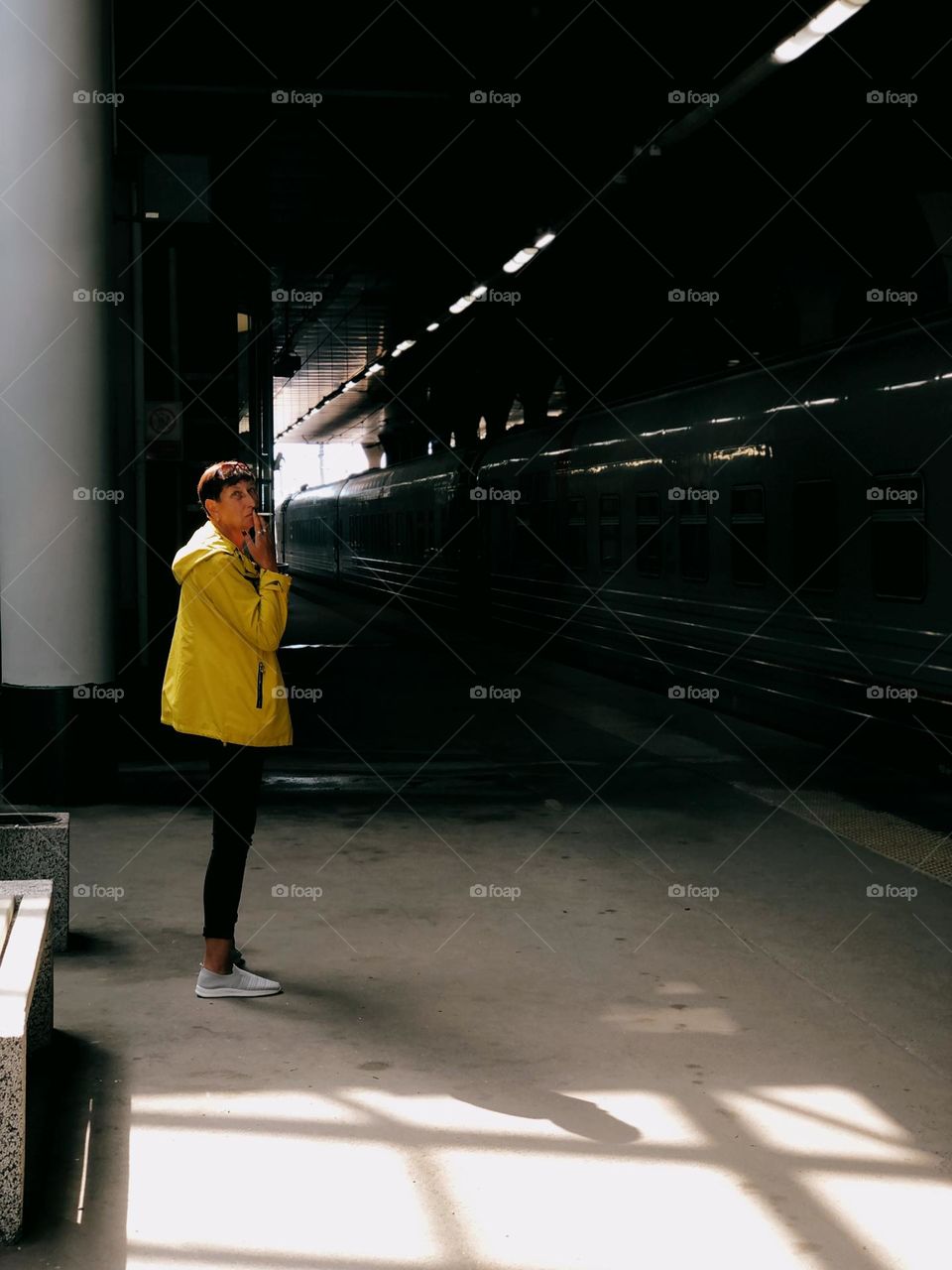 Adult woman with short hair wearing yellow jacket standing in a train station near a train, smoking.